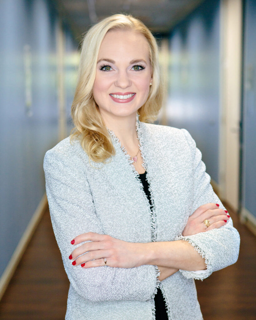 Dr Hadley Stevens Smith, a blonde, stands in a blue hallway, arms crossed, smiling at the camera